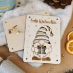 Gravierte Holzkarte mit Gnom und «Frohe Weihnachten», daneben Rückseite und goldene Rundkopfklammern, Orangenscheiben