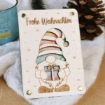 Weihnachts-Gnom Holzkarte mit bunten Geldscheinen hinter der Zipfelmütze, Tannzapfen und blaue Tasse dahinter