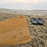 Bambus-Frühstücksbrett mit Lasergravur «in Gedanken Barfuss am Meer», Wellen und Flip-Flops, im Sand am Strand, dahinter ein Paar Flip-Flops – Geschenk für Meer- und Camperfans