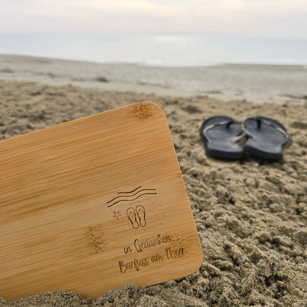 Bambus-Frühstücksbrett mit Lasergravur «in Gedanken Barfuss am Meer», Wellen und Flip-Flops, im Sand am Strand, dahinter ein Paar Flip-Flops – Geschenk für Meer- und Camperfans