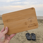 Hand hält graviertes Bambus-Brettchen «in Gedanken Barfuss am Meer» am Sandstrand, im Hintergrund Meer und Flip-Flops – kleines Geschenk für Strandliebhaber