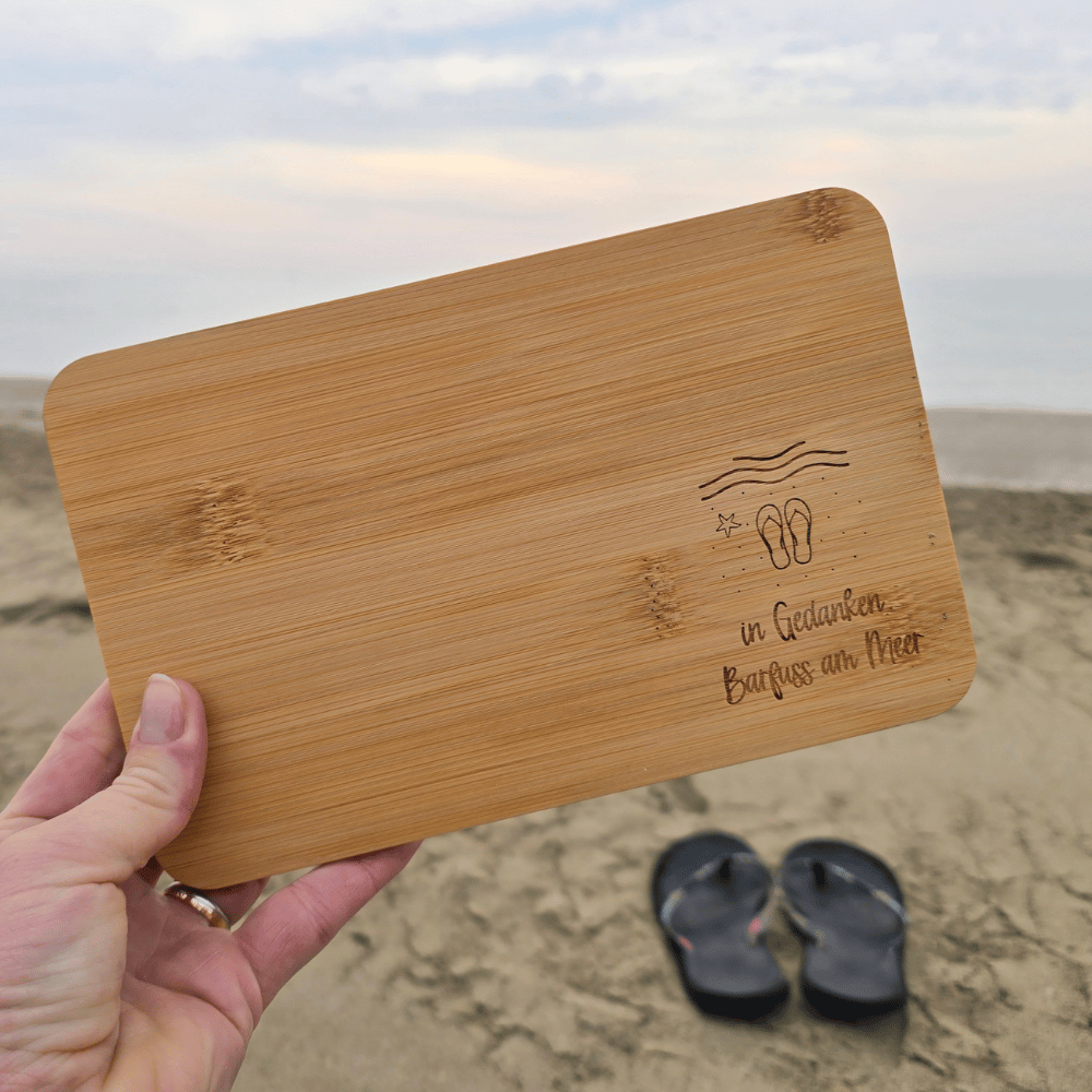 Hand hält graviertes Bambus-Brettchen «in Gedanken Barfuss am Meer» am Sandstrand, im Hintergrund Meer und Flip-Flops – kleines Geschenk für Strandliebhaber