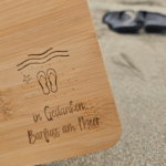 Detail der Lasergravur mit Wellen, Seestern, Flip-Flops und Spruch «in Gedanken Barfuss am Meer» auf Bambus-Schneidbrett, Flip-Flops unscharf im Sand
